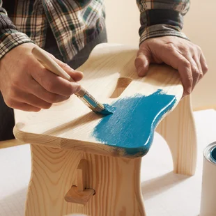 Möbel - Maler, der einen handgefertigten Holzhocker mit einem Pinsel mit einer blauen Farbe lackiert